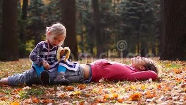 美丽的妈妈和可爱的孩子躺在秋天公园的草地上。 女儿坐在妈妈身上吃香蕉，妈妈望着天空